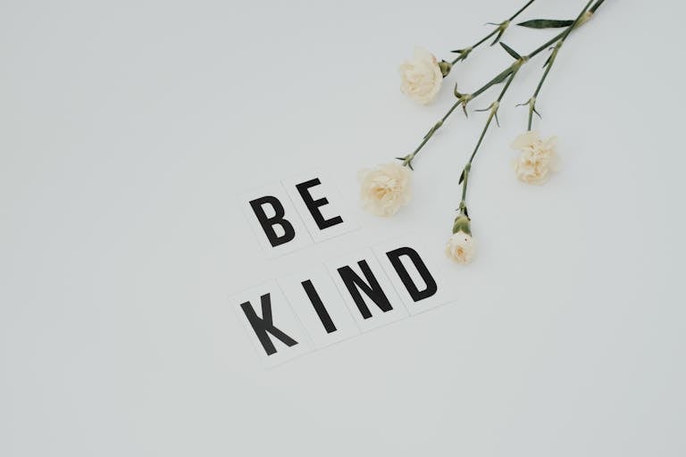 Minimalist image of 'Be Kind' text with white flowers on a light background, promoting kindness.