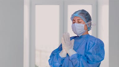 A healthcare worker in protective gear with praying hands, indoors.