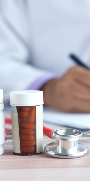 Prescription pill bottle and stethoscope on a desk with a clinician in a white coat writing in the background.