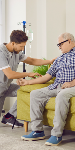 Nurse starts an IV infusion in an older man’s arm while he sits on a couch, with an IV drip stand in the background.